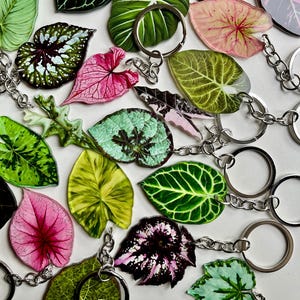 May include: A selection of leaf-shaped keyrings in shades of green, pink, and black. Each keyring showcases a detailed leaf design and is connected to a silver keyring and chain. The keyrings are displayed on a white surface.
