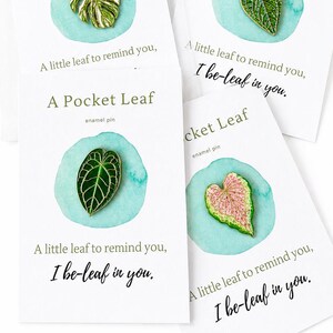 May include: Four enamel pins shaped like various leaves, including a monstera and heart-shaped varieties in green and pink, each mounted on a white card labeled A Pocket Leaf with the text A little leaf to remind you, I be-leaf in you.