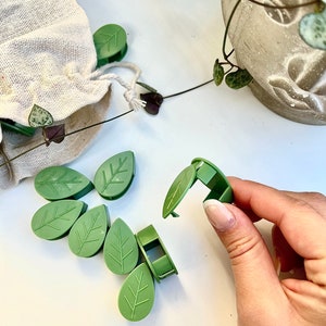 May include: Green leaf-shaped plant clips. A hand holds one open, revealing the clip mechanism. Several more clips are scattered on a white surface, alongside a small drawstring bag and a potted plant. These clips are designed to support and train climbing plants.