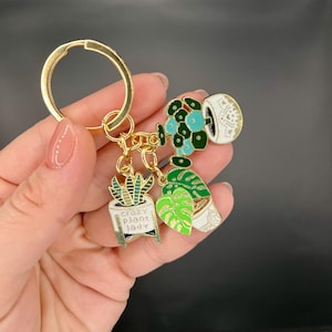 May include: Gold-coloured keyring with three plant-themed charms. The charms include a succulent, a Monstera leaf, and a plant with blue flowers. The succulent charm has the text "crazy plant lady". The charms are in white, green, and gold.