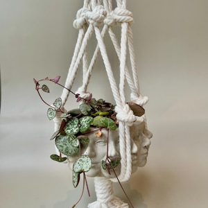 May include: A white macramé plant hanger with a grey face planter. The planter holds a string of hearts plant with green and purple leaves. The planter has three faces and is suspended by a knotted rope.