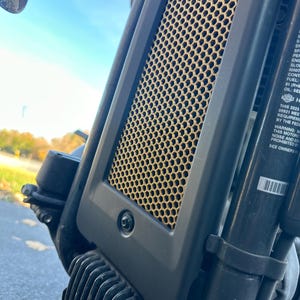 May include: Close-up of a black motorcycle part with a gold honeycomb-shaped metal grill. The grill is attached to a black plastic housing with a small black screw visible in the center. The text on the sticker reads: "THIS 2023 05921 MEET THE REQUIREMENTS BY THE FEDERAL WARNING: MOTORCYCLE NOISE AND EMISSIONS PROHIBITED SEE OWNERS MANUAL"