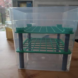 May include: A clear plastic rectangular storage container featuring an internal two-tier shelving system. The shelving consists of two green plastic grid platforms supported by four grey cylindrical pillars, creating a stacked structure inside the bin.