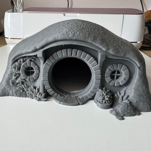 May include: A grey 3D-printed model of a hobbit hole. The structure features a round doorway, two circular windows, and a textured roof. Decorative elements include small plants and a pot. The model is designed to resemble a dwelling.