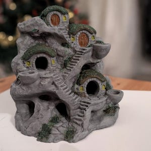 May include: A grey miniature fantasy village sculpture with multiple hobbit-hole-style dwellings. The dwellings have green thatched roofs, small windows, and wooden doors. Miniature staircases and decorative bowls are also present.