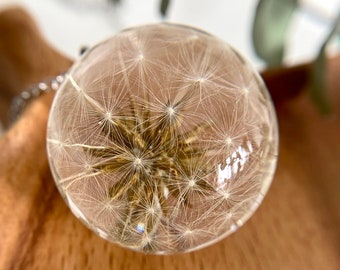 May include: A clear glass pendant with a delicate dandelion flower preserved inside. The flower is white with a brown center and the seeds are visible. The pendant is on a wooden surface.