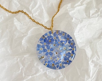 May include: A gold chain necklace with a round pendant featuring a pressed forget-me-not flower design.