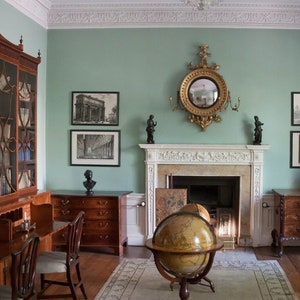 May include: A vintage-style room with a fireplace, a globe, and a large wooden bookcase. The walls are painted a light green color and there are several framed prints on the walls. The room is decorated with antique furniture and accessories.