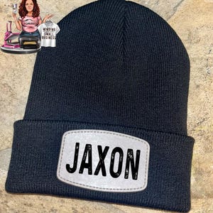 May include: A black knit beanie with a rectangular patch that reads "JAXON" in bold, black letters. The patch has a white background and a stitched border. The beanie is made of a ribbed knit material.