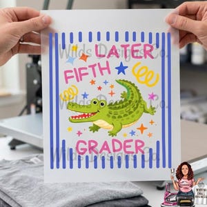 Pu&ograve; includere: Un foglio di trasferimento termico con un alligatore verde dei cartoni animati e le parole "LATER FIFTH GRADER" in rosa e blu. Il design include stelle e spirali, con strisce verticali blu sui lati.