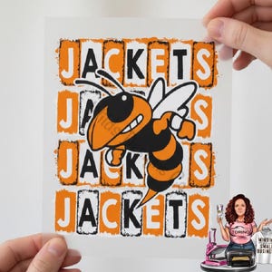 May include: A white transfer sheet with the word "JACKETS" in orange and black block letters. A cartoon bee in black and orange is in the center. The image is held by two hands.