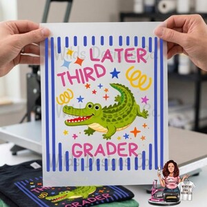 Pu&ograve; includere: Un foglio di trasferimento a sublimazione bianco con la scritta "Later Third Grader" e un alligatore dei cartoni animati. Il disegno include strisce verticali blu, stelle e coriandoli. Il foglio &egrave; tenuto in mano.