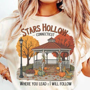May include: Cream-colored t-shirt featuring a fall-themed graphic. The design includes a gazebo, pumpkins, and autumn trees, with the text "STARS HOLLOW CONNECTICUT" and "WHERE YOU LEAD I WILL FOLLOW".