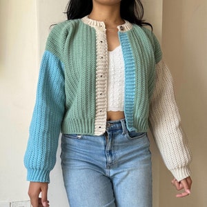 Puede incluir: Un cárdigan corto con un diseño de bloques de color verde, azul y crema. El cárdigan tiene cierre de botones y mangas largas.