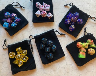 May include: Nine black velvet bags with different colored dice inside. The dice are in various shapes and sizes, with numbers and symbols on them. The bags are all closed and have a drawstring.