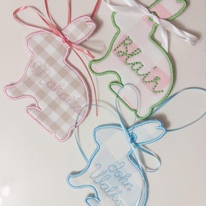 Op de afbeelding: Drie geborduurde konijnvormige labels met namen. Eén is roze en bruin geruit met een roze lint, een andere is wit en roze met een groene omtrek en wit lint, en de laatste is blauw en wit met een blauw lint.
