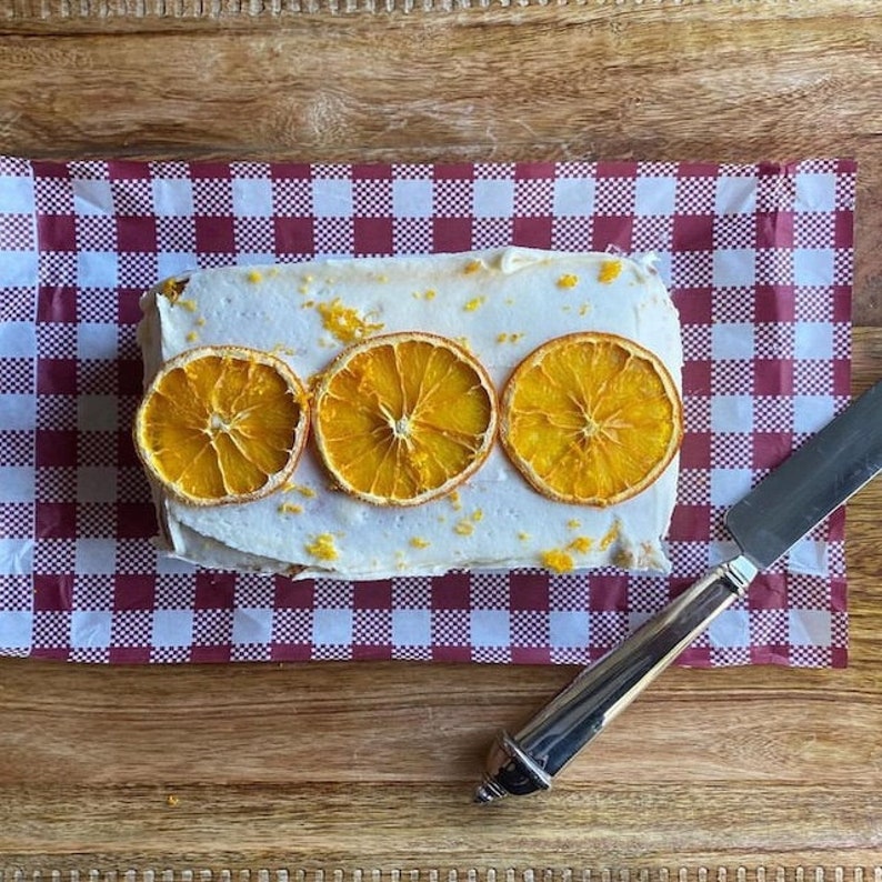 May include: A loaf cake with white icing and three orange slices on top. The cake is sitting on a red and white gingham paper.