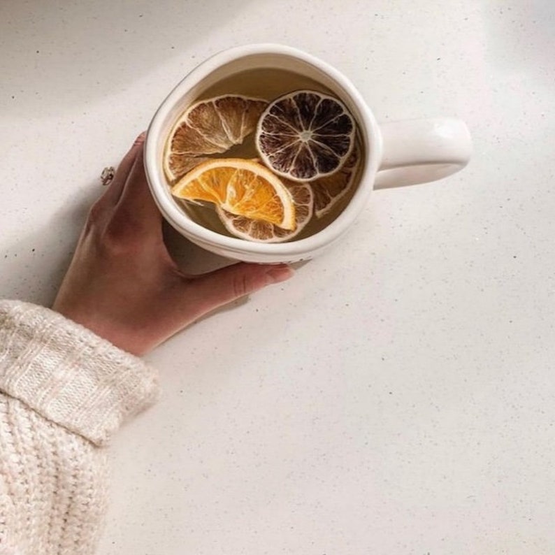 May include: A white ceramic mug filled with a light yellow liquid and dried orange slices. The mug has a rounded handle.