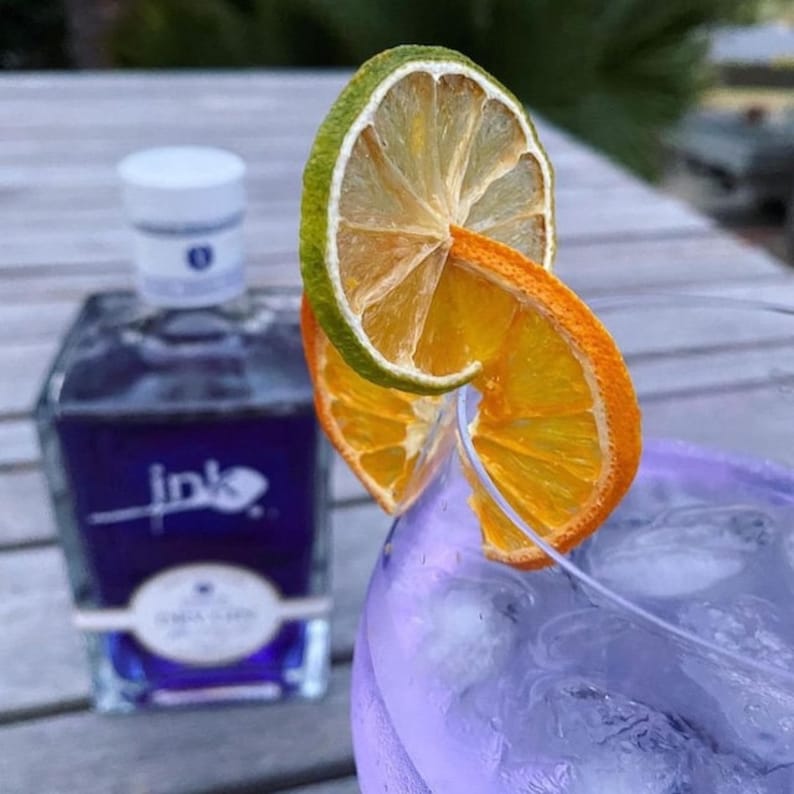 May include: A purple bottle of Ink Gin with a white label and the word "Ink" in a stylised font. The bottle is next to a glass of purple liquid with ice and a garnish of dried orange and lime slices.