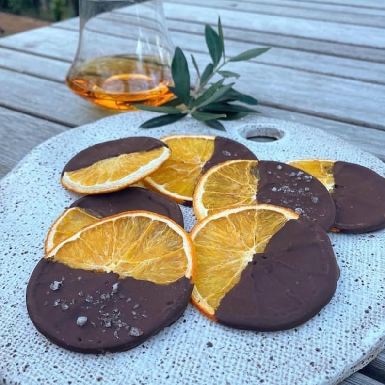 May include: A white ceramic serving platter with several orange slices dipped in dark chocolate. The chocolate is topped with a sprinkle of sea salt.