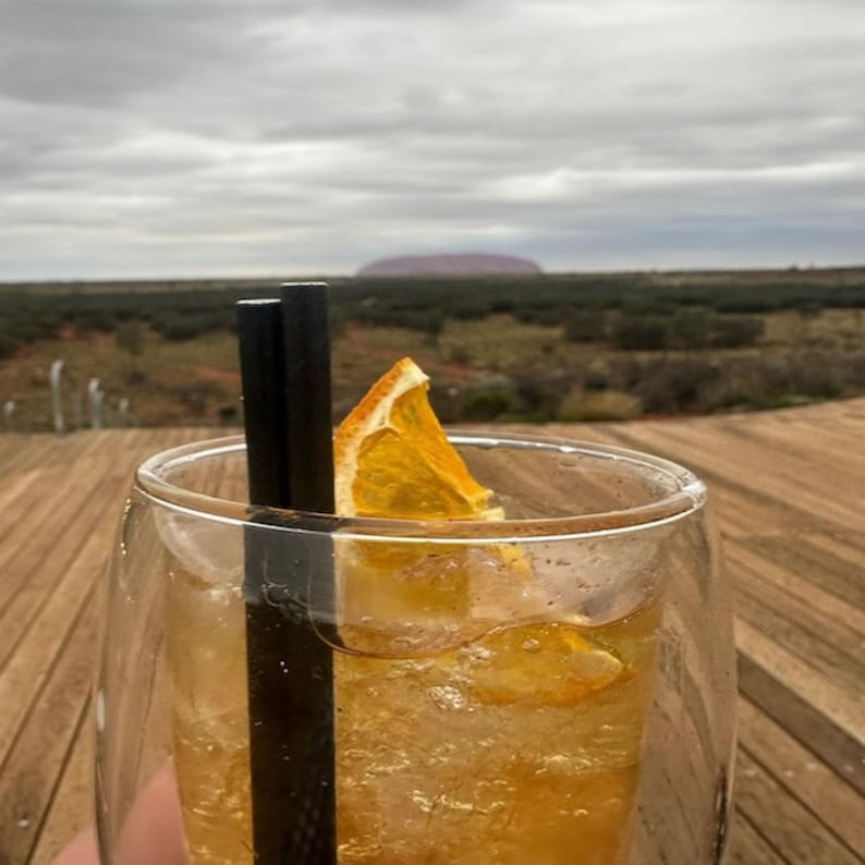 May include: A glass of iced tea with an orange slice garnish and two black straws. The drink is on a wooden table with a view of a large rock formation in the distance.