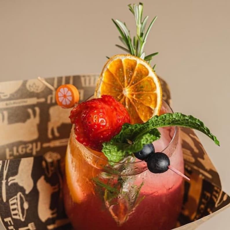 May include: A pink cocktail garnished with a strawberry, orange slice, rosemary sprig, and blueberries. The drink is in a clear glass and is sitting on a brown paper coaster with a pattern of cows and the word "fresh" written on it.