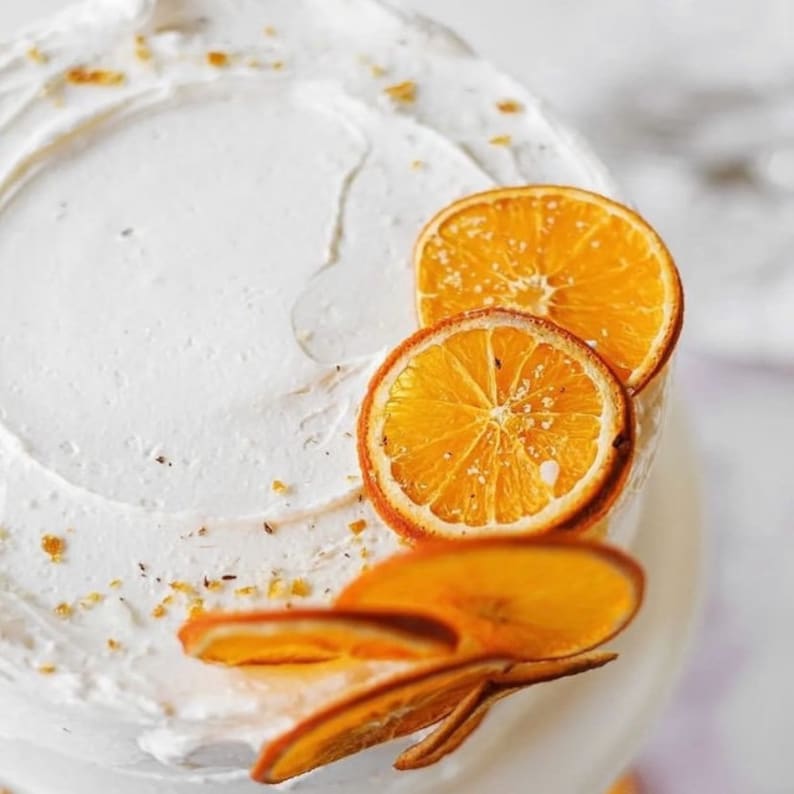 May include: A white cake with a swirl of frosting and orange slices on top. The cake is decorated with small, white sprinkles.
