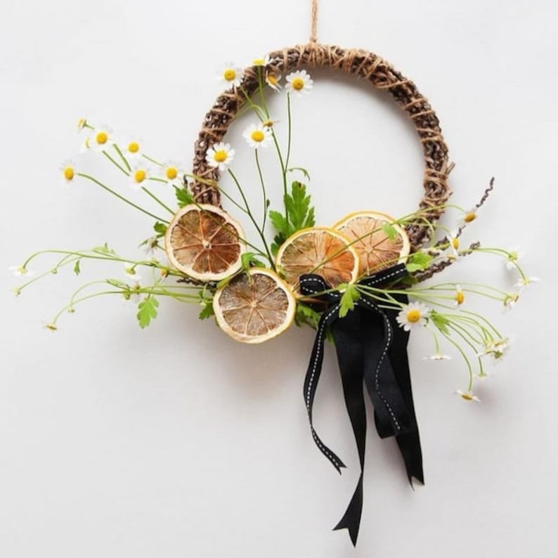 May include: A wreath made of natural twine with dried lemon slices and white daisies. A black ribbon is tied at the bottom of the wreath.