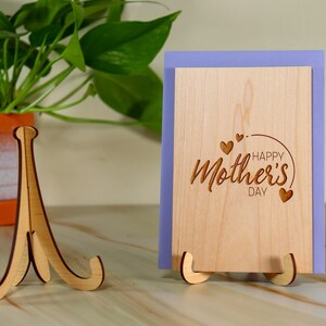 May include: A wooden card stand with a light brown finish holds a card with the text "Happy Mother's Day" engraved on it. The card is surrounded by a purple border.