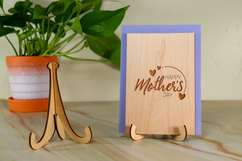 May include: A wooden card stand with a light brown finish holds a card with the text "Happy Mother's Day" engraved on it. The card is surrounded by a purple border.