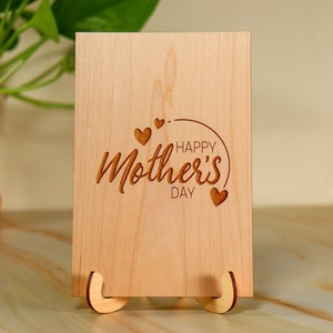 May include: A wooden card with the text "Happy Mother's Day" engraved on it. The card is decorated with small hearts and is standing on a wooden easel.