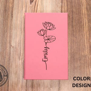 May include: A rectangular pink notebook featuring a black line art illustration of a water lily flower and lily pads. The name Emily is written in a black cursive script below the floral design. The text COLOR: PINK and DESIGN: JULY are visible.
