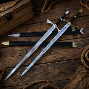 May include: Two long swords with patterned metal blades and black grips. One sword features a silver-toned pommel and crossguard, while the other has a gold-toned pommel and crossguard. Two black scabbards with metal tips are positioned behind the swords.