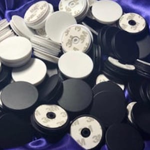 May include: A collection of white and black round phone grips with adhesive backing. The grips are in various stages of assembly, with some showing the 3M adhesive pads. The image is a close-up shot, highlighting the product's design and functionality.
