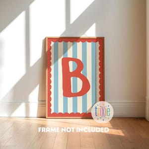 Pode incluir: Uma impressão emoldurada com a letra "B" em vermelho, sobre um fundo de listras verticais azuis e creme. A moldura tem uma borda vermelha ondulada. O texto "FRAME NOT INCLUDED" está na parte inferior. A impressão está numa sala com chão de madeira.