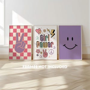 May include: Three framed art prints with a retro theme. The first print has a pink and white chequered background with a peace sign. The second print says "Girl Power" with hearts, flowers, and a peace sign. The third print is a purple background with a smiley face.