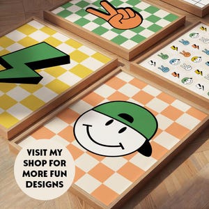 May include: A collection of framed art prints with a retro theme. Each print features a unique design, including a peace sign, a lightning bolt, and a smiley face wearing a cap, all set against a checkered background. The text "VISIT MY SHOP FOR MORE FUN DESIGNS" is also visible.
