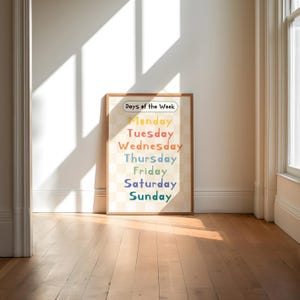 May include: A framed print displaying the days of the week in a rainbow of colours. The text is set against a checkered background. The words "Days of the Week" are at the top. The frame is a light brown colour.