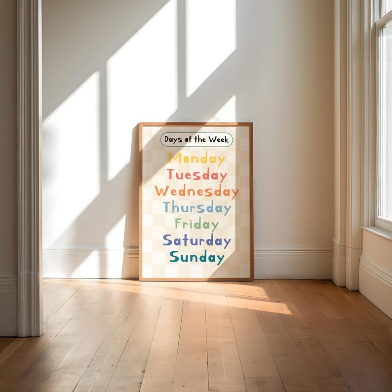 May include: A framed print displaying the days of the week in a rainbow of colours. The text is set against a checkered background. The words "Days of the Week" are at the top. The frame is a light brown colour.