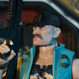 Può includere: Action figure di un uomo calvo con i baffi, che indossa un berretto nero e un gilet turchese. La figura ha un tatuaggio dettagliato sul petto e tiene in mano un fucile nero. Una piccola granata è attaccata al gilet.