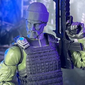 May include: A detailed action figure of a soldier in olive green and black tactical gear, holding a black rifle. The figure wears a gray beanie and a tactical vest. The figure has a serious expression.