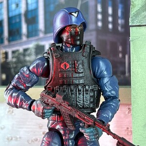 Può includere: Action figure blu di un soldato con elmetto, copertura per il viso nera e giubbotto tattico. La figura tiene in mano un fucile con mirino rosso. Il giubbotto ha un simbolo di cobra rosso.