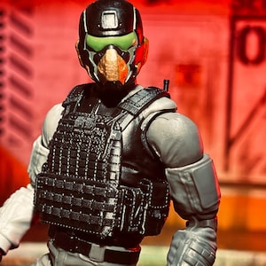 May include: A detailed action figure of a soldier in tactical gear. The figure wears a black helmet with green visor, a black tactical vest, and gray uniform. The background is a blurred red.