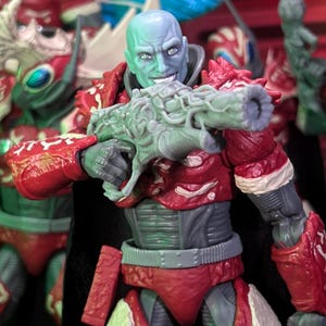 Peut inclure: Figurine d'action détaillée d'un extraterrestre humanoïde avec une tête bleue et verte, une armure rouge et une arme grise. La figurine sourit et tient l'arme, avec d'autres figurines similaires en arrière-plan. L'armure de la figurine est riche en détails.