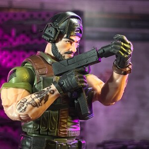 May include: A male action figure with a dark beard, wearing a black cap, tactical vest, and headset. The figure has a black tattoo on the right forearm, holds a black submachine gun, and wears black gloves. The figure is dressed in olive green.