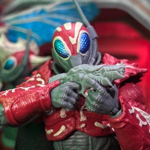 Peut inclure: Figurine d'action détaillée d'un guerrier futuriste. La figurine est vêtue d'une armure rouge et grise, tenant une arme grise. Le casque présente un design rouge avec des yeux bleus et verts. L'esthétique générale suggère un thème de science-fiction ou de fantaisie.