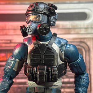 Puede incluir: Figura de acción detallada de un soldado con equipo táctico. La figura es principalmente azul y gris, con casco, máscara de gas y chaleco táctico con bolsillos. La figura tiene articulaciones y detalles realistas.