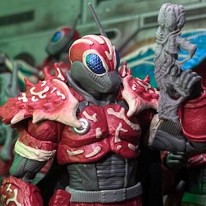 Peut inclure: Figurine d'action détaillée d'un guerrier futuriste en armure rouge et grise. La figurine a un casque distinctif en forme d'insecte avec des yeux bleus et tient une arme grise. L'armure présente des détails complexes en forme d'os.