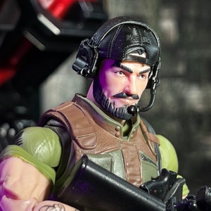 Puede incluir: Figura de acción de un hombre con gorra negra, auriculares y chaleco marrón sobre una camisa verde. La figura tiene barba y bigote. La figura sostiene un arma negra.