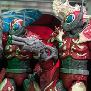 Peut inclure: Gros plan sur deux figurines d'action. Les figurines sont vêtues d'une armure rouge et verte avec des détails complexes. Elles ont des casques avec des visières bleues et tiennent des armes grises d'aspect futuriste. Les figurines sont posées dans une posture dynamique, suggérant l'action.